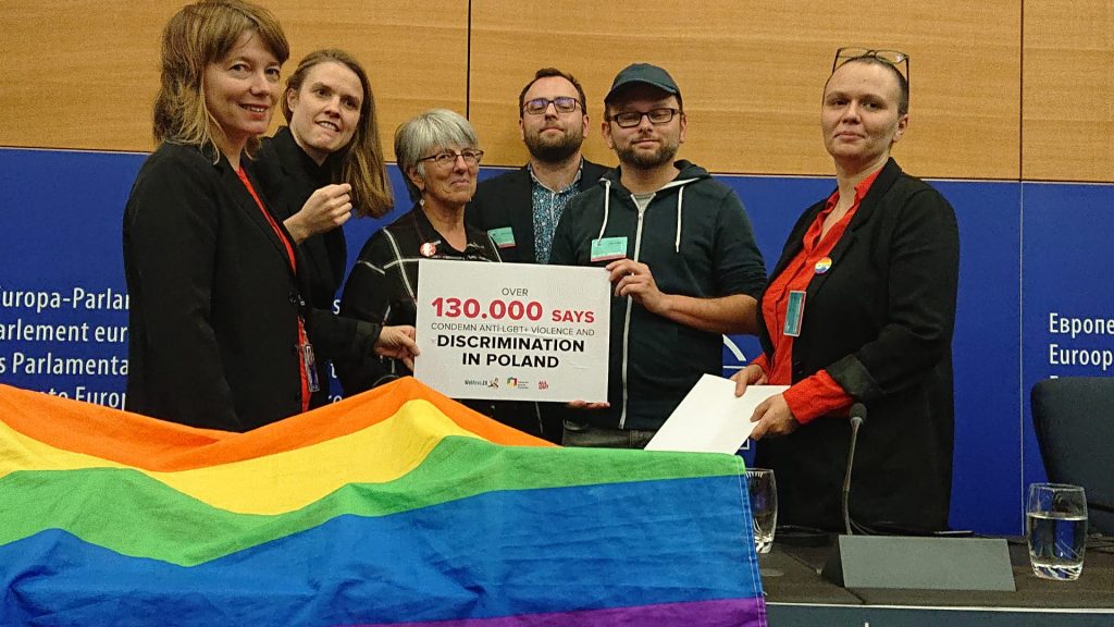 Zdjęcie przedstawia grupę ludzi trzymających tabliczkę z napisem: 130.000 podpisów przeciwko uchwałom anty-LGBT (po angielsku) i tęczową flagą